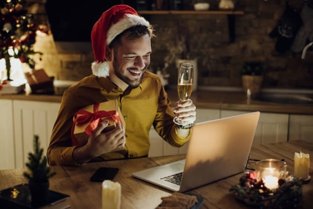 Young happy man having video call over laptop and toasting with Champagne while celebrating Christmas alone at home.